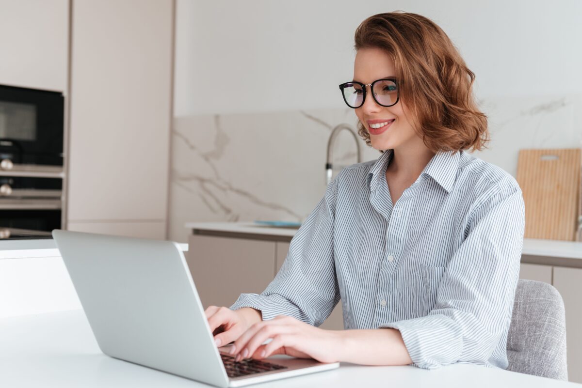 Elegant smiling woman in glasses and striped shirt using computer computer while sitting at the table in the kitchen min